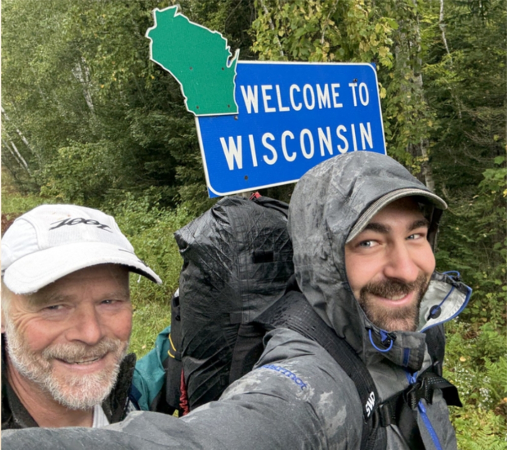 Matt Ostrander in a white baseball cap and Colin P. Tingo in a rain jacket with his hood up smiling in front of a "Welcome to Wisconsin" sign in a forested area.